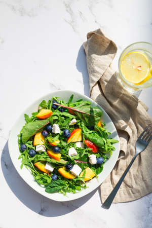Fresh homemade salad with nectarines, blueberries, arugula, spinach and feta cheese on white marble background with hard shadows. Healthy vegetarian food. Top view.の写真素材