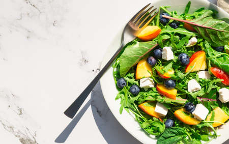 Fresh homemade salad with nectarines, blueberries, arugula, spinach and feta cheese on white marble background with hard shadows. Healthy vegetarian food. Top view. Space for text.の写真素材