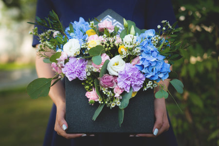 Image of flowers arrangement with hydrangea, white eustoma, spray yellow and violet roses, pink carnations, waxflowers. A girl is holding bright mixed flowers bouquet in black envelope boxの写真素材