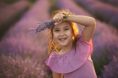 Beautiful Little girl is holding small bouquet in the lavender field. Close-up portrait of cute kid girl in the purple dress at sunsetの写真素材