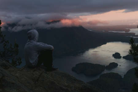 Man sitting on the rim with campfire above coastal view with islands and forests in cloudy weather from top view while sunsetの写真素材