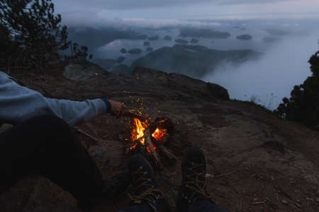 Man sitting on cliff with campfire above coastal view on bay in the eveningの写真素材