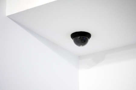 Dome CCTV in black. A hemisphere camera is mounted on the ceiling. The ceiling and walls of the room are plain white. Internal, external, hidden video surveillance.の写真素材