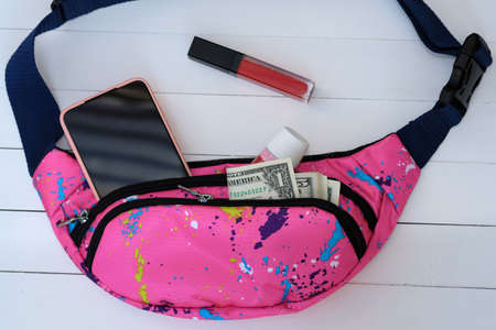 Banana bag with phone, lipstick, cream and dollar bills. Waist bag pink color on a white wood background. A small fabric bag with two compartments for storing your phone, cosmetics and money.の写真素材