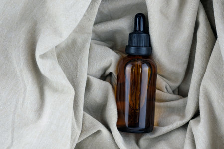 Glass bottle with a pipette on a background of linen cloth, top view. A brown bottle of essential...