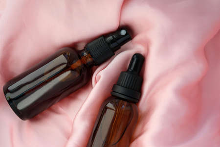 Cosmetic bottles for serum and tonic. Two glass bottles on pink silk top view. Brown glass containers with a pipette and a spray bottle. Nutritious cream, gel, body lotion with protein of silk.の写真素材