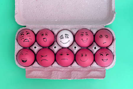 Painted red eggs in a cardboard tray top view. Eggs painted with emotional faces and a white egg. Ten eggs scattered on a green background top view. White crow among others. positive attitude, unlikeの写真素材