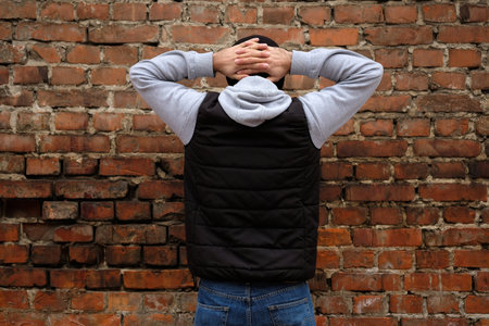 The guy stands facing a brick wall with his hands clasped behind his head. A guy in jeans and a black puffy tank top with his back to the camera. Arrested man with hands behind his head.の写真素材