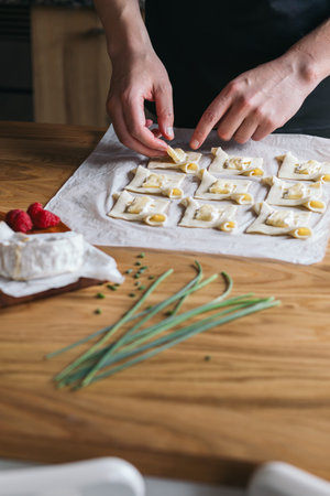 A person is placing pieces of camembert cheese onto puff pastry squares on a parchment-lined tray....