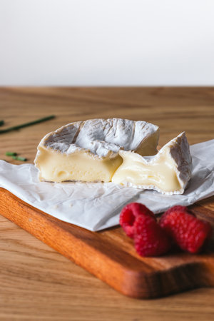 Close up of a wheel of soft camembert cheese with a trimmed wedge and fresh raspberries placed on a wooden cutting board.の写真素材