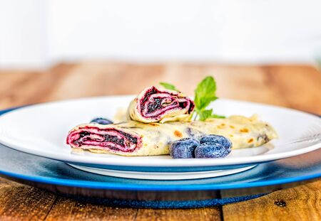 Lateral view of crepe with blueberry jam filling cut in half and decorated with mint leaves and frozen blueberries on white and blue plates.の写真素材