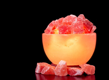 Turned on Himalayan pink salt lamp carved as a bowl on a black table with a black background.の写真素材