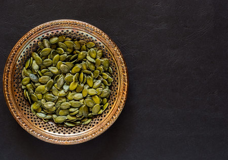 Raw pumpkin seeds in an antique meral bowl on a dark textured backgroundの写真素材