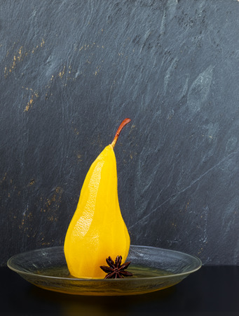 Single pear poached in sugar and saffron syrup spiced with star anise on a glass plate with a gray slate backgroundの写真素材