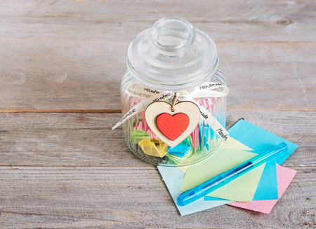 Glass jar with handmade wooden hearts decorations and ribbon near a stack of colored papers and a blue pen.の写真素材