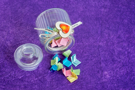 Toppled over glass jar with handmade wooden hearts decorations and ribbon full of colorful folded paper slips.の写真素材