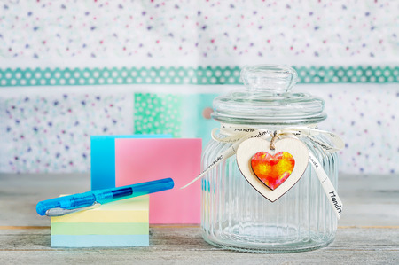 Glass jar with handmade wooden hearts decorations and ribbon near a stack of colored papers and a blue pen.の写真素材