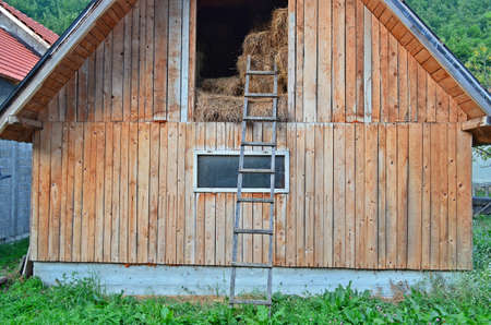 A mountain village wooden sawn timber barn with hay bales inside, a ladder in front and forest in the backgroundの写真素材
