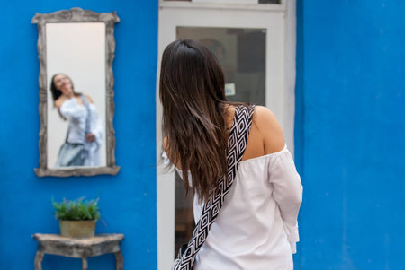 Beautiful woman on white dress looking at herself on a mirror on the colorful streets of the...