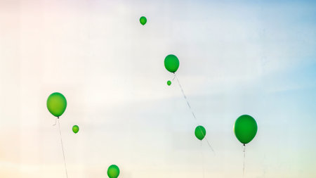 Vibrant green balloons ascending into pale blue and white sky background for celebrationの素材