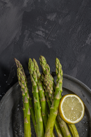 Bundle of young raw uncooked organic green asparagus with sliced lemon and sea salt over black texture background. Top view. Healthy eatingの写真素材