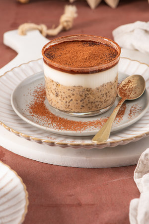 Tiramisu chia pudding in a glass, topped with cocoa powder, served on a white plate with a spoon, creating a visually appealing dessert presentationの写真素材