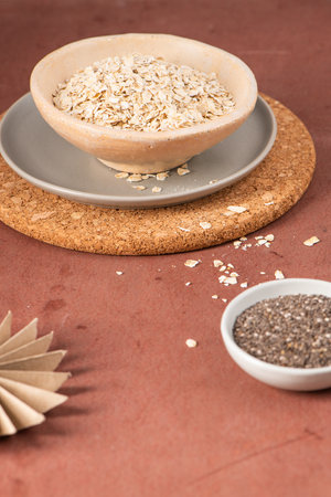 Bowl of oats on a cork mat with scattered oats and a small bowl of chia seeds on a brown surfaceの写真素材