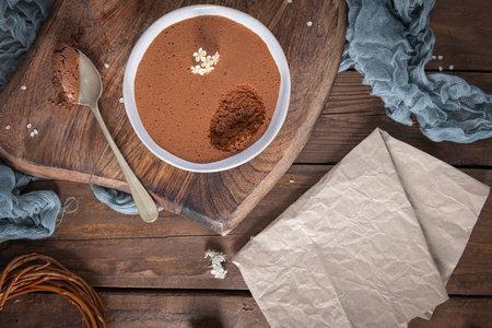 A white bowl filled with rich chocolate mousse, a spoonful partially removed, is presented on a wooden board with a silver spoon and delicate white flowers. Top-Down Viewの写真素材
