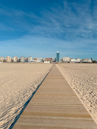 A wooden boardwalk stretches across a vast sandy beach, leading towards a cityscape with tall buildings against a clear blue sky in Figueira da Foz, Portugalの写真素材