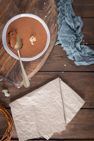 A creamy chocolate mousse in a white dish is presented on a wooden board with a golden spoon,...