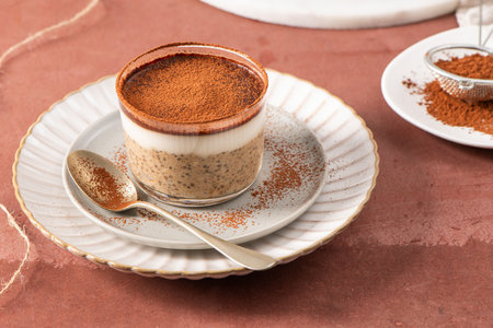 Tiramisu chia pudding in a glass cup on a white plate with a spoon, garnished with cocoa powder, set on a brown surface with a sieve and cocoa powder in the backgroundの写真素材