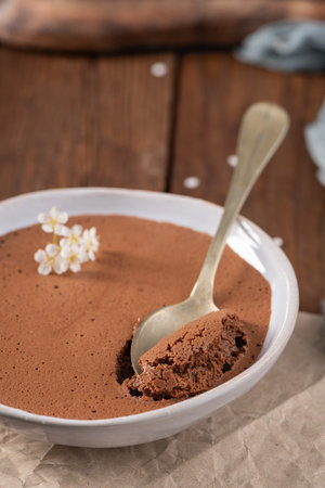 Silky chocolate mousse in a white bowl, a spoonful being lifted, decorated with tiny white flowers, presented on crumpled parchment paper and a rustic wooden tableの写真素材