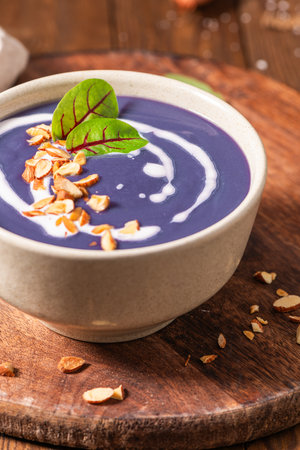 A bowl of creamy purple beet soup topped with a swirl of cream, toasted almonds, and fresh herbs.の写真素材