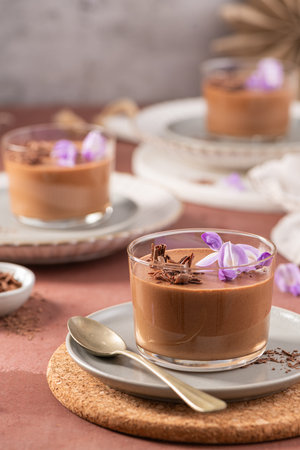 Multiple servings of smooth chocolate mousse in glasses, elegantly garnished with purple flowers and chocolate shavings on a rustic surface.の写真素材