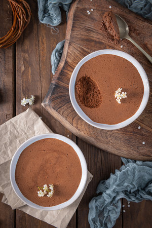 Creamy chocolate mousse in a white bowl, elegantly presented on a wooden board with a silver spoon and delicate white flowersの写真素材