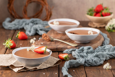 Creamy chocolate mousse in a white bowl, garnished with delicate white flowers and a fresh strawberry, set on rustic parchment paper and a warm wooden surfaceの写真素材