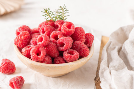 A rustic ceramic bowl filled with plump, fresh raspberries sits on a white table surrounded by...