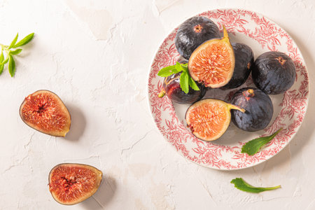 A close-up, top-down view of whole and halved figs on a decorative plate with fresh leaves and two fig halves scattered on a white backgroundの写真素材