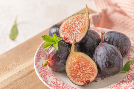 A close-up of whole and halved ripe figs on a decorative plate with fresh leaves A wooden board, a pink napkin, and a white backgroundの写真素材