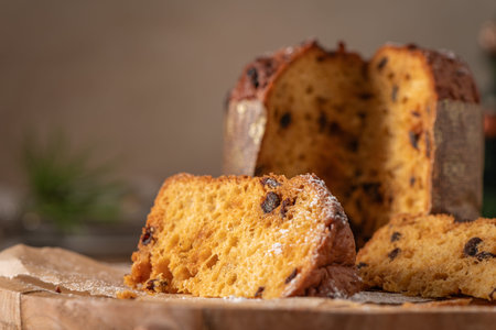 A warm close-up of a generous slice of traditional Italian Panettone with raisins and powdered sugar, set on a wooden board with the whole cake blurred in the backgroundの写真素材