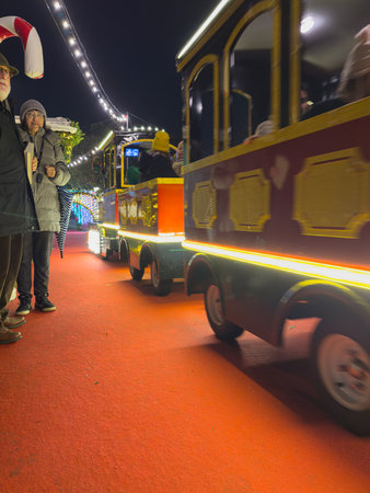 Espinho, Portugal - December 6, 2025. A brightly lit miniature train travels along a vibrant red carpet pathway past illuminated wooden Christmas market stalls and an older couple at night in Espinho Portugal, capturing the festive atmosphereのeditorial素材