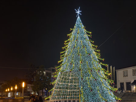 Santa Maria da Feira, Portugal - December 6, 2025. A dazzling, massive Christmas tree illuminated with thousands of white and yellow lights and red ornaments stands tall at night. A few people gather near the base, capturing photos on the cobblestone squaのeditorial素材