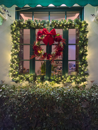 Santa Maria da Feira, Portugal - December 6, 2025. A close-up, centered view of a dark green window pane framed by thick green garland and bright string lights, featuring a lavish wreath with red roses, silver ornaments, and a large red velvet bowのeditorial素材