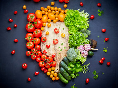 Colorful vegetables composition with red and yellow tomatoes, cucumbers, broccoli, greens. Wooden cutting board next to which lie small tomatoes and parsley on a black background. Concept of cooking healthy food for vegans and vegetarians. Top view.の写真素材