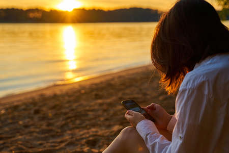 Young millennial girl sitting on the sandy shore at sunset and looking at the screen of her smartphone. Last days of the summer holidays. Alone traveling woman using her Mobile Phoneの写真素材