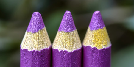 A close up of three purple and yellow colored pencils with a blurred green background outsideの素材