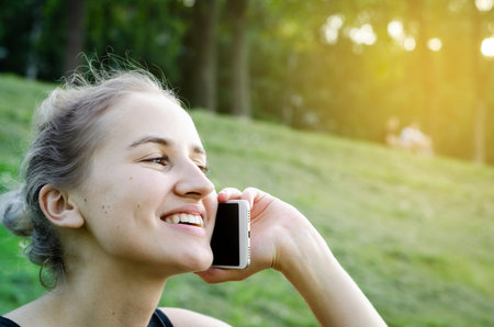 Girl talking on the phone and laughing. Communication through distance. Sitting in the Park. The blonde in sports . The glare of the sun. Photo.の写真素材