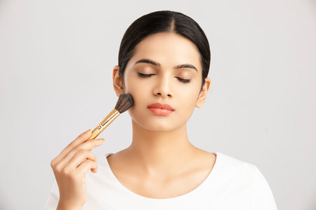 Beautiful young woman with makeup brush on light background. Space for text