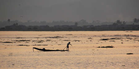A fisherman is standing on his boat, and fishing with a net  The sun is setting and the lake lights up in it s golden rays   In the background is a village with palm trees の写真素材