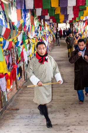 A man in Thimpu, whearing the traditional bhutanese costume is walking over a bhutanes bridge, that is covered completely with prayer flagsのeditorial素材